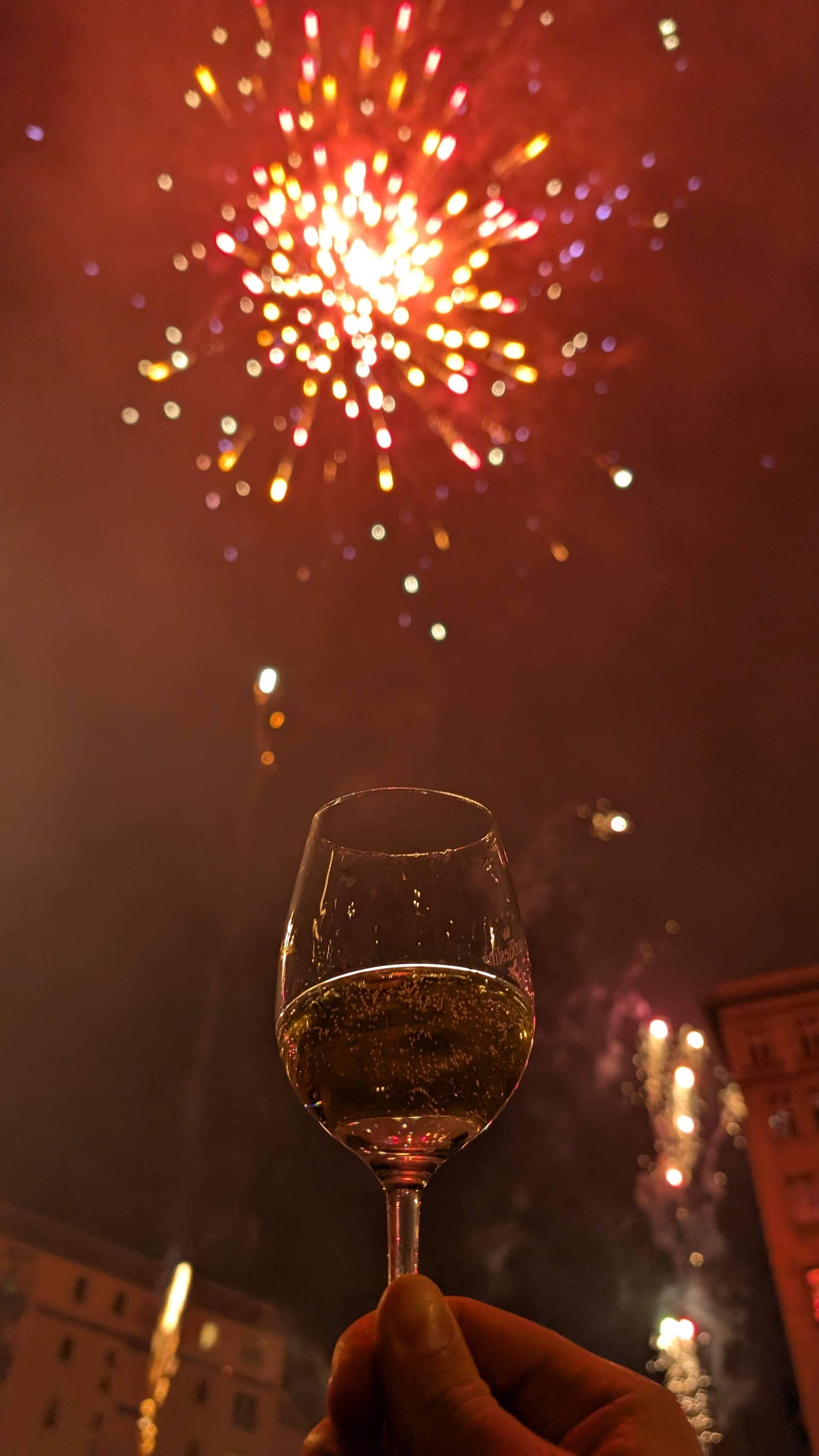 Eine Hand hält ein Weinglas und im Hintergrund ist Feuerwerk zu erkennen