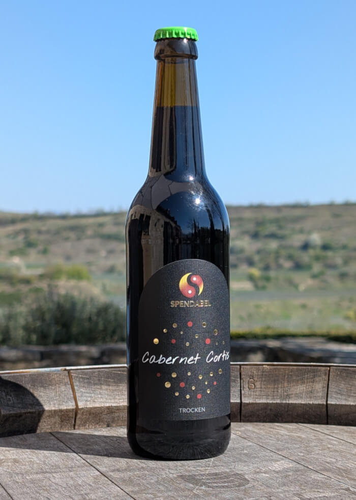 0,5 Liter Flasche SPENDABEL Rotwein Cabernet Cortis trocken auf einem Weinfass mit Weinbergen im Hintergrund