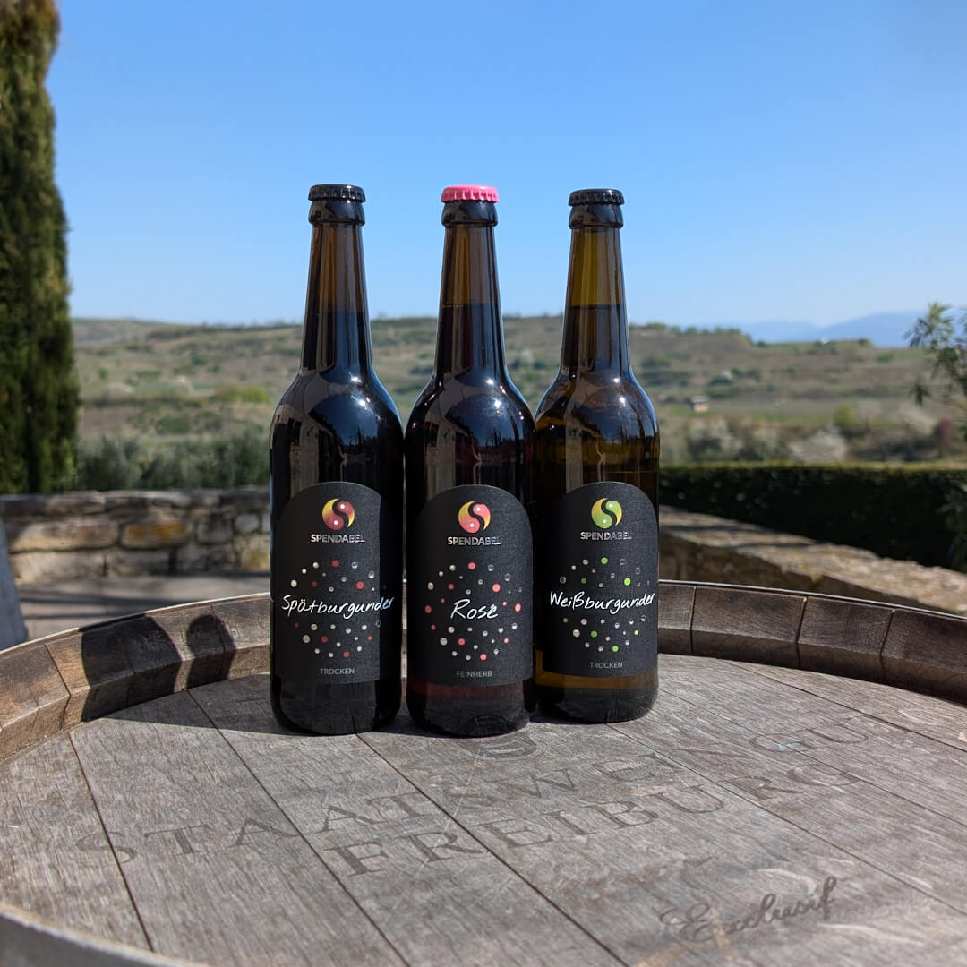 Jeweils einer Flasche der SPENDABEL Weine, Weißburgunder trocken, Rosé feinherb und Spätburgunder trocken auf einem Weinfass aus Holz. Im Hintergrund sind Weinberge.