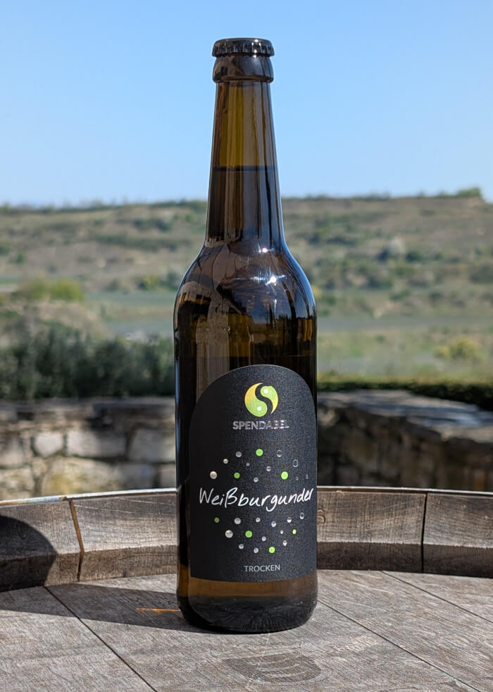 0,5 Liter Flasche SPENDABEL Weißwein Weißburgunder trocken auf einem Weinfass mit Weinbergen im Hintergrund