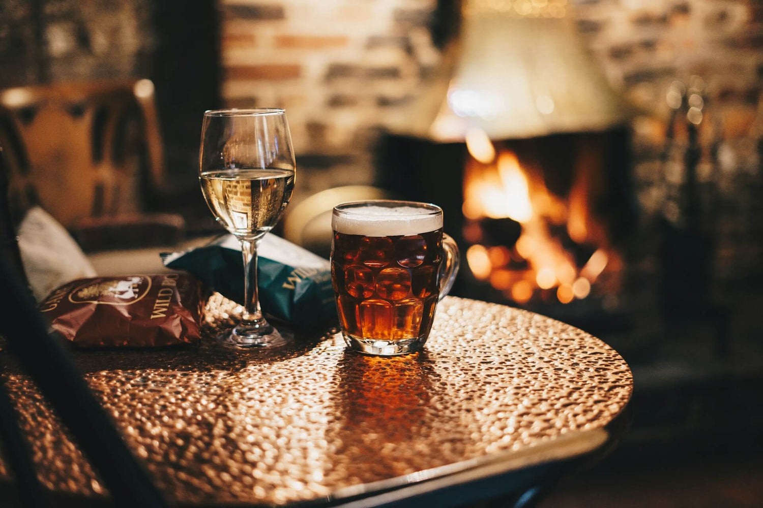 Ein Wein und ein Bierglas stehen auf einem Tisch und im Hintergrund ist ein Kamin mit offenem Feuer zu sehen.